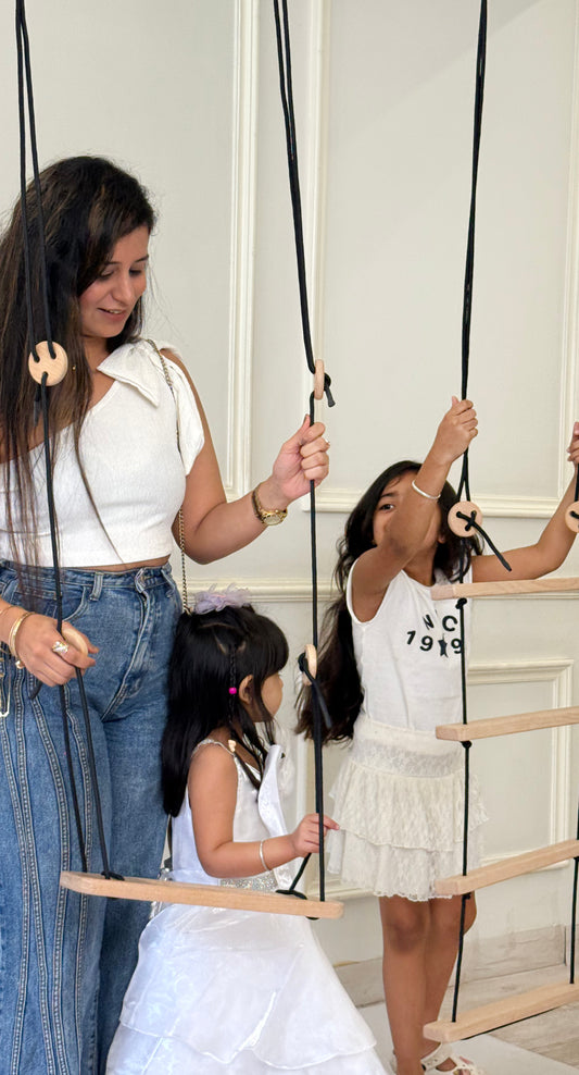Woman and two young girls playing with a wooden swing and climbing toy indoors.
